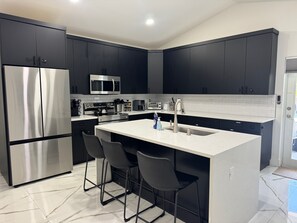 A modern kitchen with dark cabinets, stainless steel appliances, a central island with bar stools, and a white countertop. - Stylish 4BR /2 Bath Oasis with Heated Pool, Hot Tub - 10 Mins to Beach/Downtown (West Palm Beach)