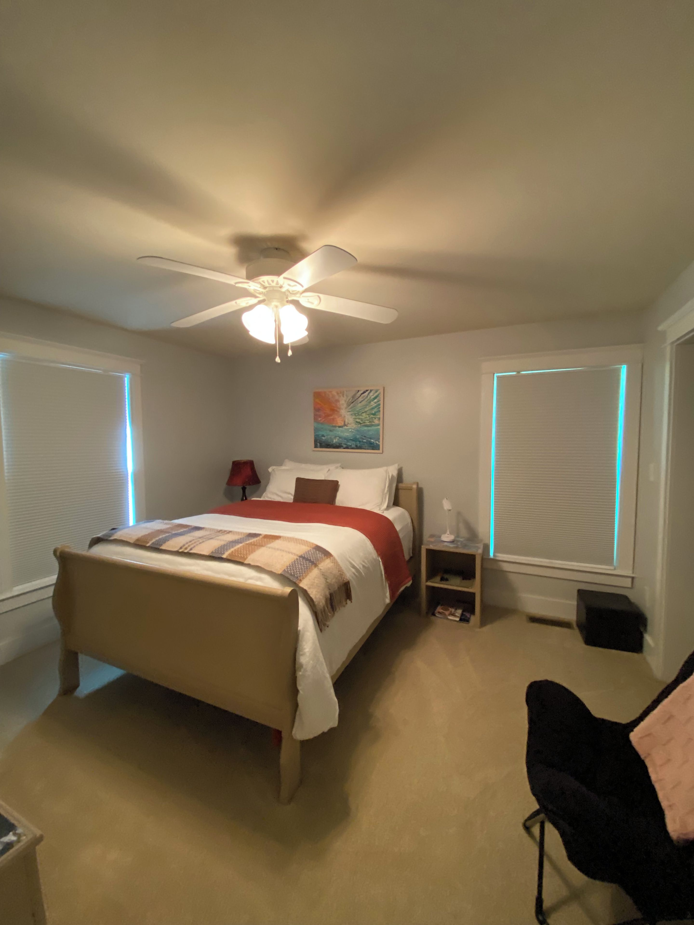 Un dormitorio con cama, ventilador de techo, mesita de noche, lámpara y ventana con persianas.