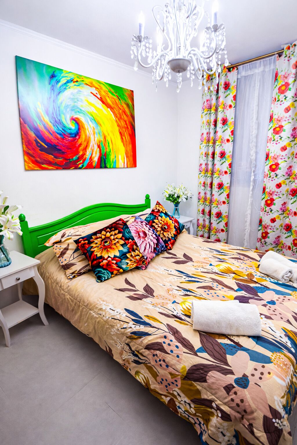 A bedroom with a green headboard, a colorful abstract painting, a floral bedspread, a white nightstand, a vase with flowers, and a chandelier.