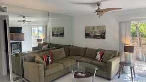 Uma sala de estar moderna com um sofá seccional, uma mesa de centro de vidro, um ventilador de teto e um espelho grande que reflete o ambiente.