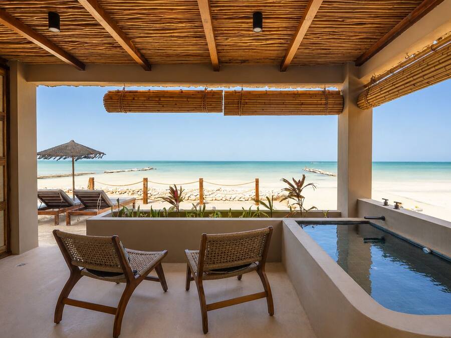 Un balcón con vista a la playa, dos sillones de mimbre, una piscina y un techo de paja.
