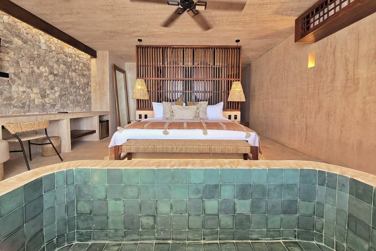 A bedroom with a large bed, a wooden headboard, a stone wall, a desk with a chair, and a bathtub with green tiles.