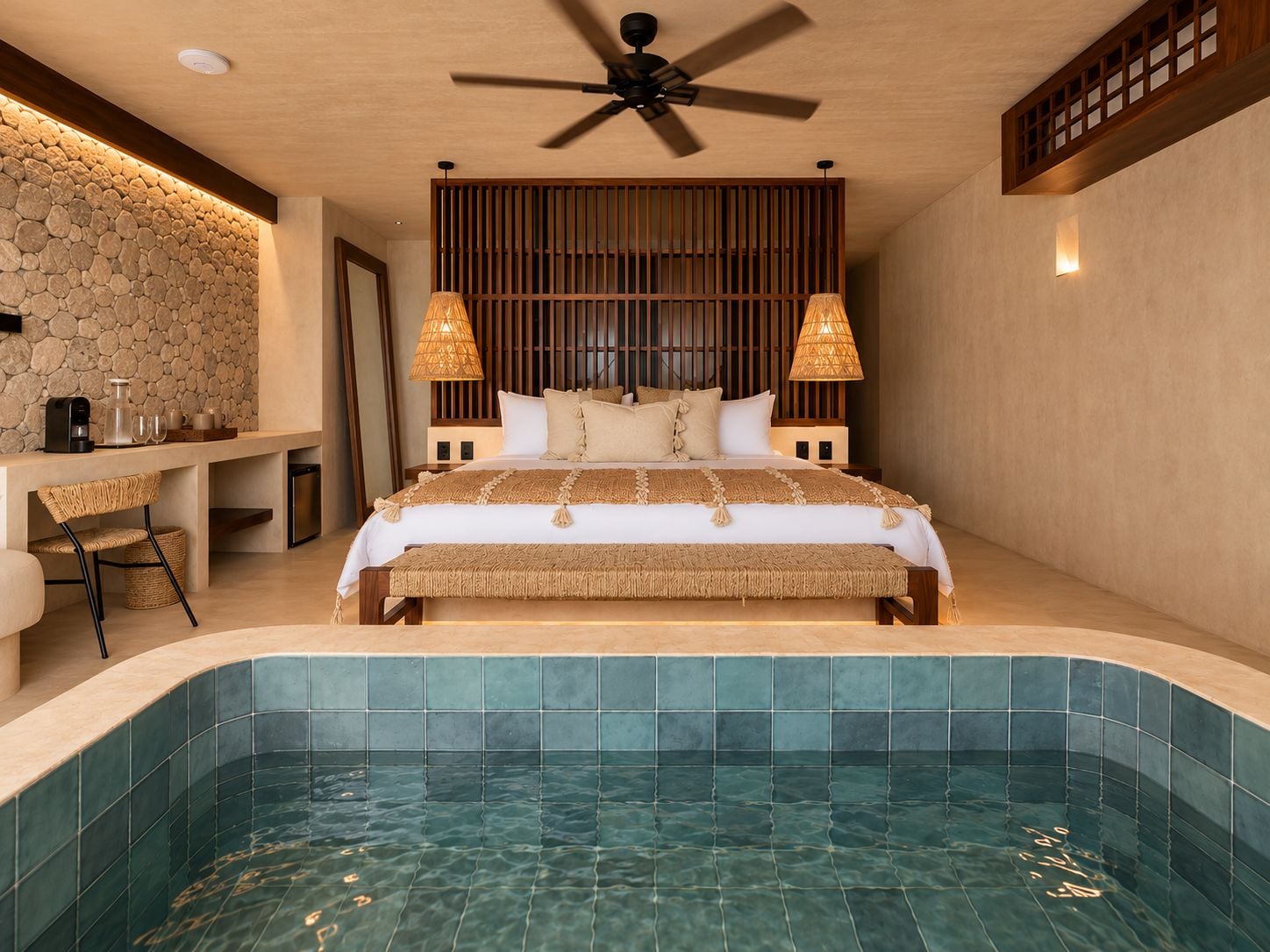 Un dormitorio con una cama grande, lámparas de noche, un cabecero de madera, una pared de piedra, un pequeño escritorio con silla y una piscina integrada.