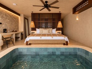 Un dormitorio con una cama grande, lámparas de noche, un cabecero de madera, una pared de piedra, un pequeño escritorio con silla y una piscina integrada.