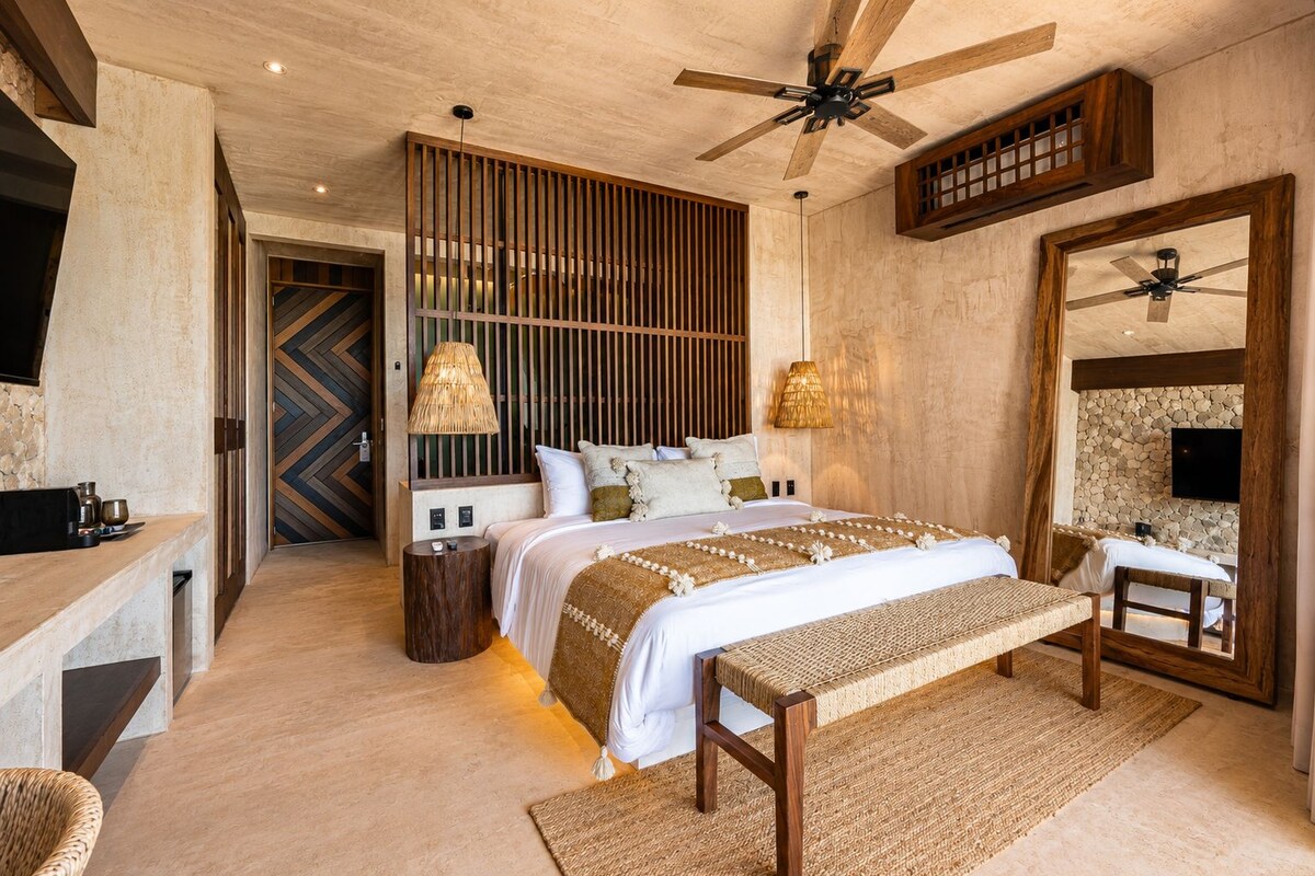 A modern bedroom with a large bed, a wooden headboard, a ceiling fan, and a stone wall.