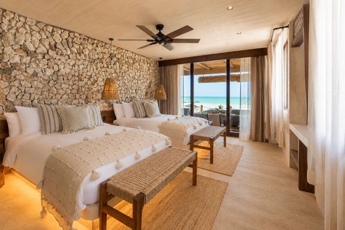 A hotel room with a stone wall, two beds, a ceiling fan, and a view of the beach.