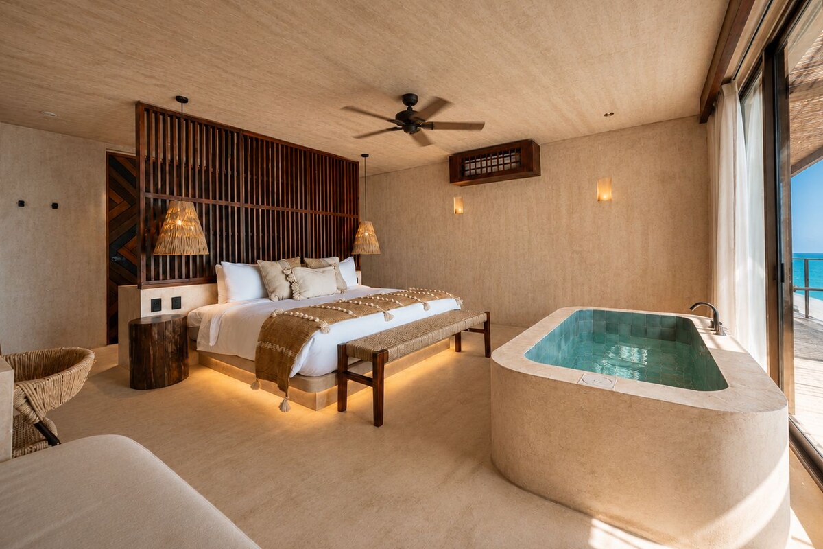 A bedroom with a large bed, a wooden headboard, a bench, a bathtub, and a view of the sea.