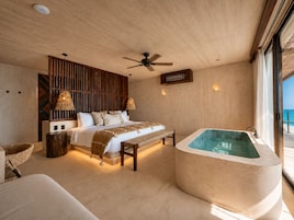 A bedroom with a large bed, a wooden headboard, a bench, a bathtub, and a view of the sea.