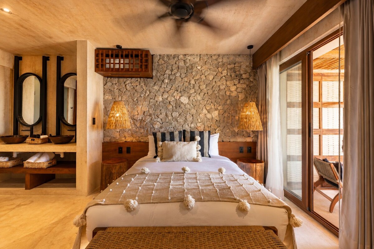 A bedroom with a stone wall, a bed with a checkered bedspread, two bedside lamps, and a large window with curtains.