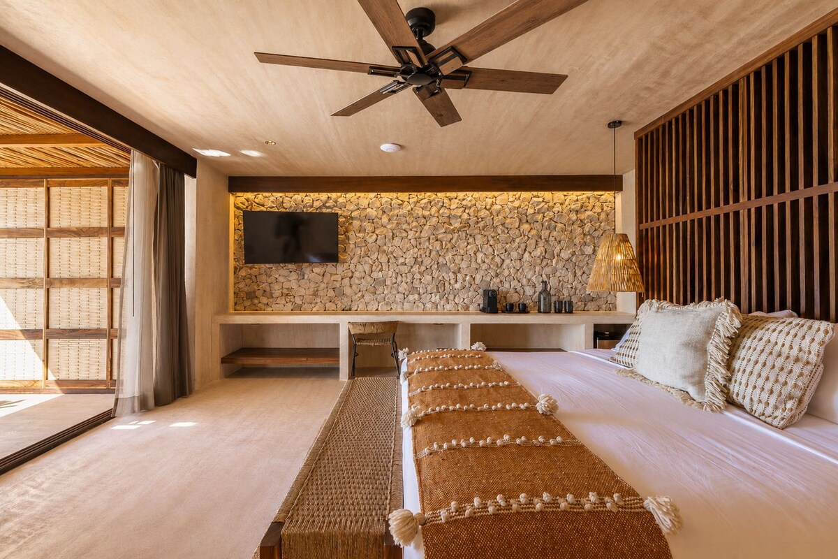 A modern bedroom with a large bed, a ceiling fan, a stone wall, and a TV mounted on the wall.