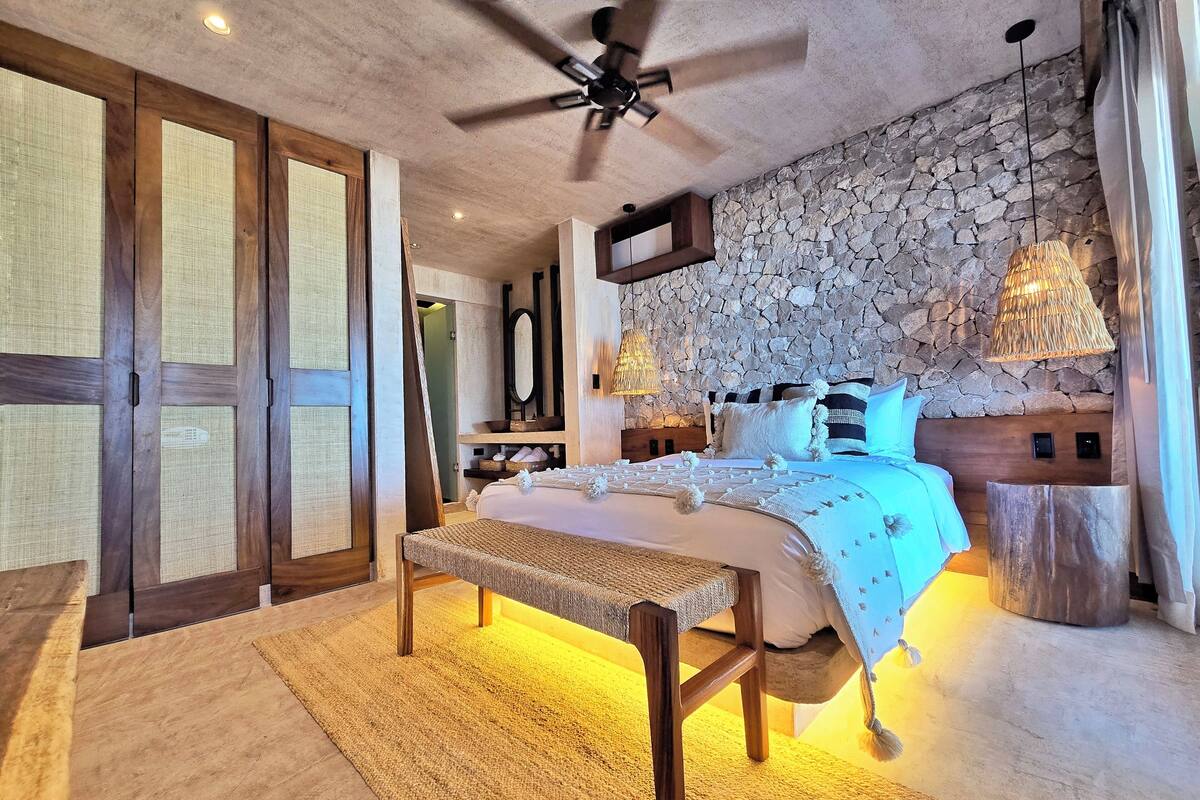 A bedroom with a stone wall, a bed with a wooden headboard, a bench, and a ceiling fan.