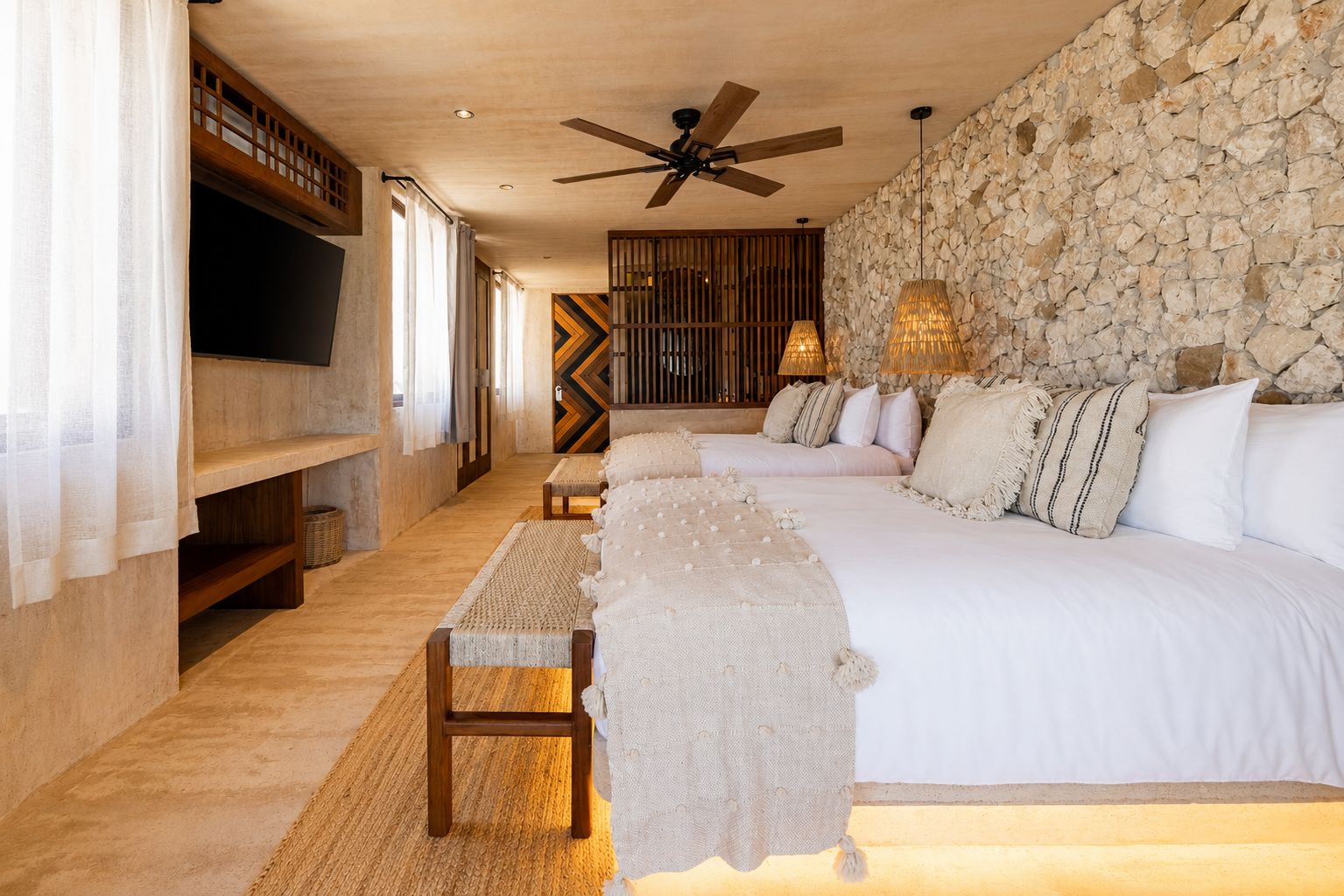 Un dormitorio con pared de piedra, ventilador de techo, televisor y cama con muchos almohadones.