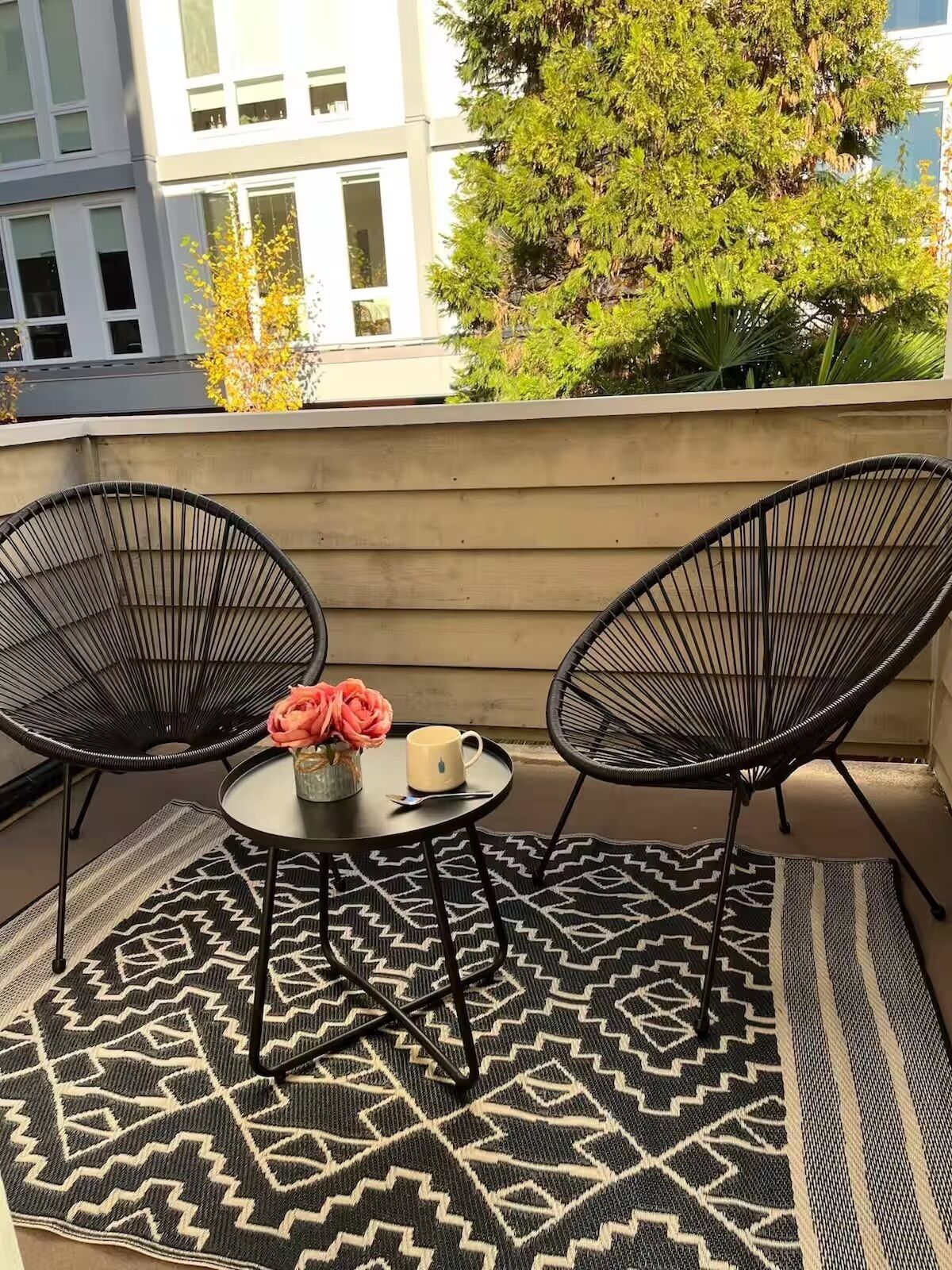 Un balcone con due sedie nere in fil di ferro, un tavolino rotondo con un vaso di fiori e una tazza, e un tappeto decorato.