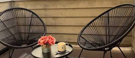Un balcone con due sedie nere in fil di ferro, un tavolino rotondo con un vaso di fiori e una tazza, e un tappeto decorato.