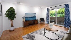 A modern living room with a flat-screen TV, wooden flooring, a large potted plant, a sofa, a coffee table, and a view through a glass door. - Spacious Roosevelt Urban Lodge + Walk to Light Rail (Seattle)
