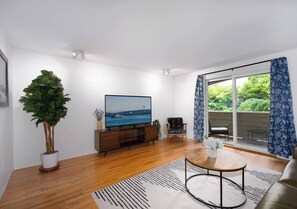 A modern living room with a flat-screen TV, wooden flooring, a large potted plant, a sofa, a coffee table, and a view through a glass door. - Spacious Roosevelt Urban Lodge + Walk to Light Rail (Seattle)