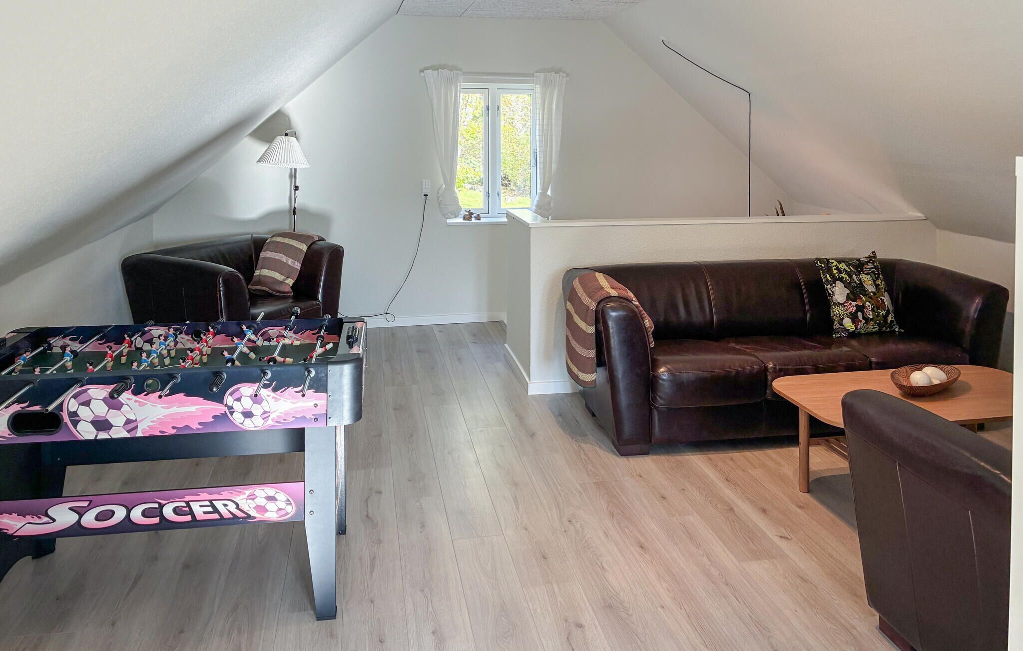 Acogedor loft con mesa de fútbol, sofá de cuero, una pequeña mesa con un bol y ventana con cortinas.