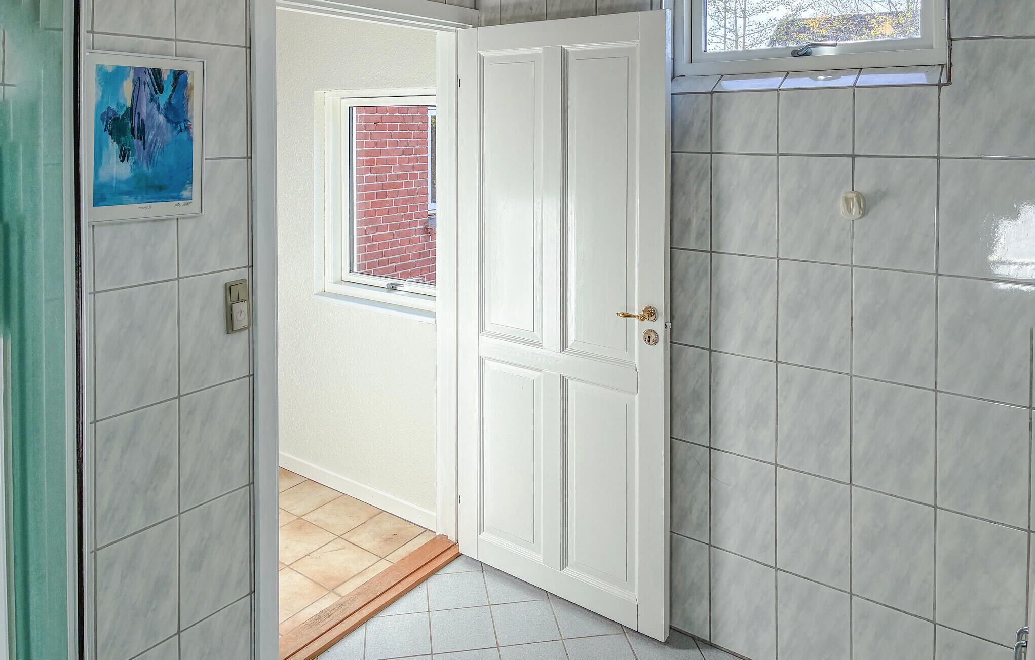 A hallway with a white door, a window with a view of a brick wall, and a tiled floor.