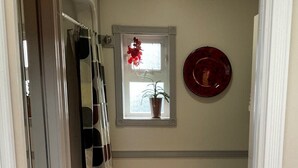 Une petite salle de bain avec une douche, des toilettes, une fenêtre ornée d’un pot de fleurs et une assiette rouge accrochée au mur.