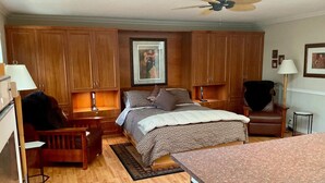 Une chambre à coucher comprenant un lit, une armoire en bois, des tables de chevet, une chaise, un bureau, un ventilateur de plafond et un tableau encadré accroché au mur.