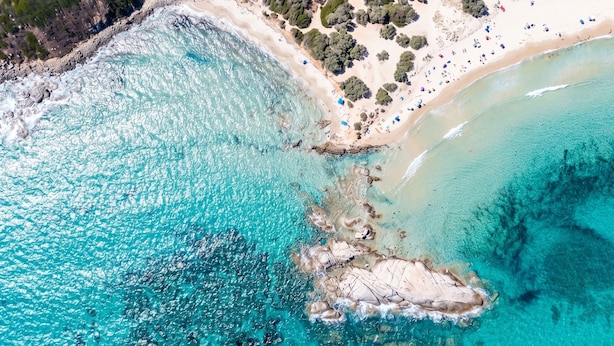 Une vue aérienne d’une plage aux eaux turquoise et limpides, avec des formations rocheuses et une étendue de sable où se trouvent des personnes et des parasols.