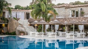 Un espace piscine agrémenté de meubles en osier blanc et de parasols, un bâtiment en pierre avec un toit en tuiles, et un escalier menant à un balcon.