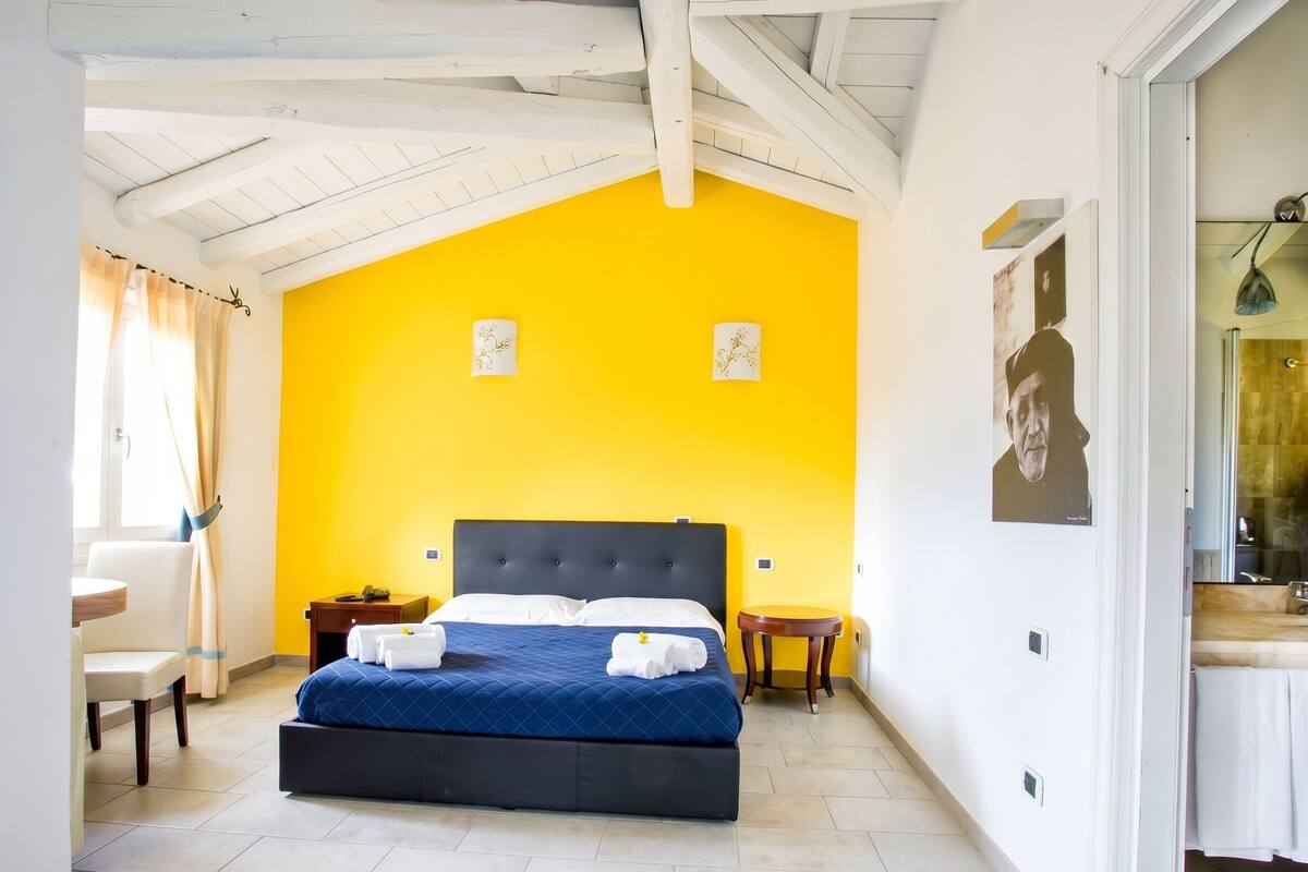 Een kamer met een bed, een klein tafeltje, een stoel en een raam met gordijnen.