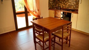 A dining area with a wooden table and chairs, a kitchen with wooden cabinets and a tiled backsplash, and a sliding glass door with curtains. - La Luna Nel Pozzo (Sciacca)