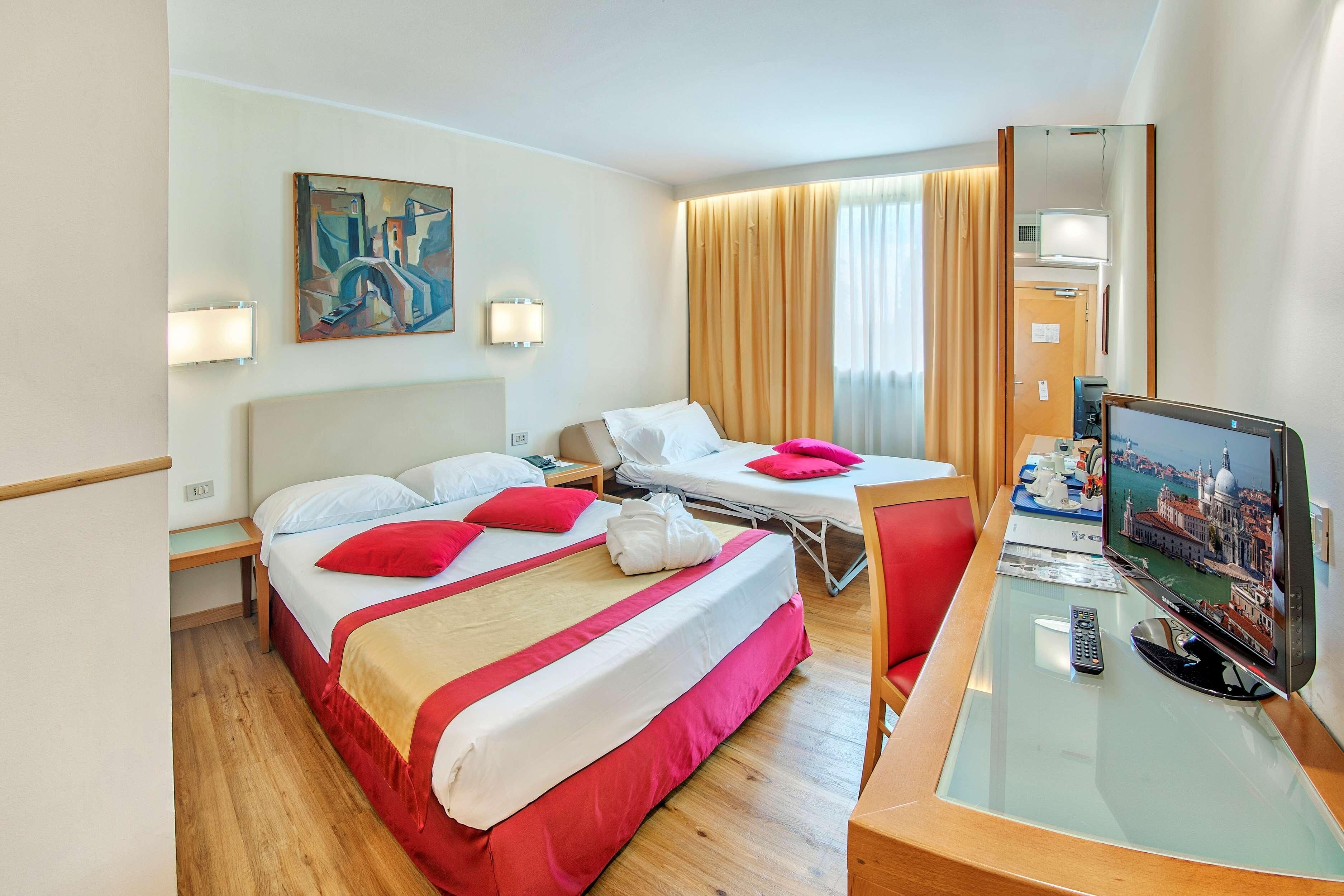 A hotel room with a bed, a desk with a computer, a TV, and a painting on the wall.