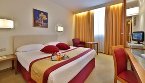 A hotel room with a bed, two red pillows, a bedside table with a lamp, a chair, a desk with a computer, and a window with curtains. - B&B Hotel Quarto D'altino (Quarto d'Altino)