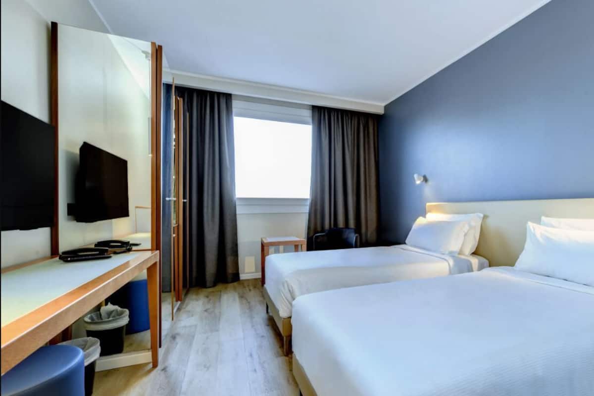 Hotelkamer met twee bedden, een bureau, een flatscreen televisie, een raam met gordijnen en een prullenbak.