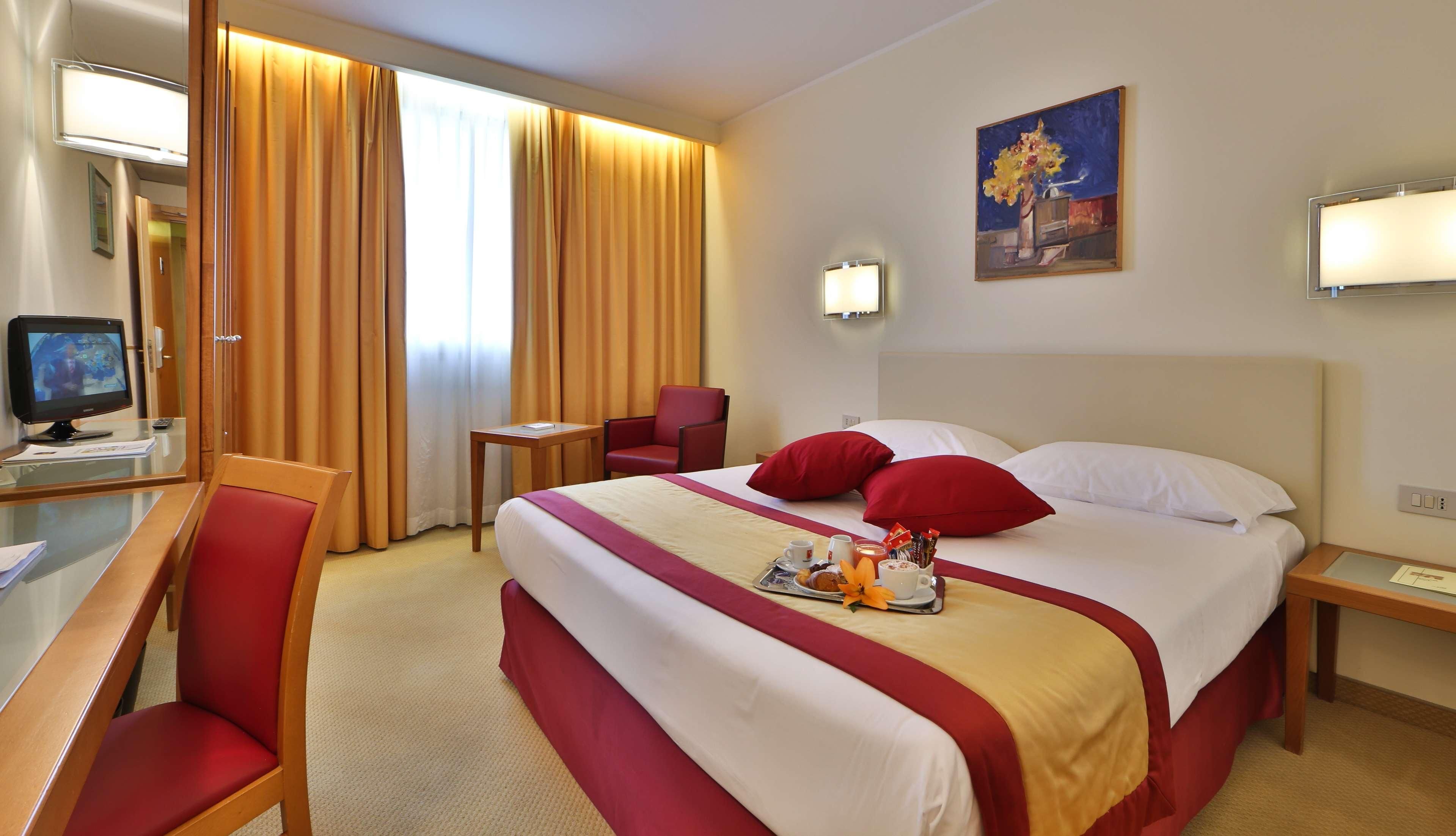 A hotel room with a large bed, red and white bedding, a desk with a computer, a chair, and a painting on the wall.