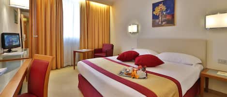 A hotel room with a large bed, red and white bedding, a desk with a computer, a chair, and a painting on the wall.