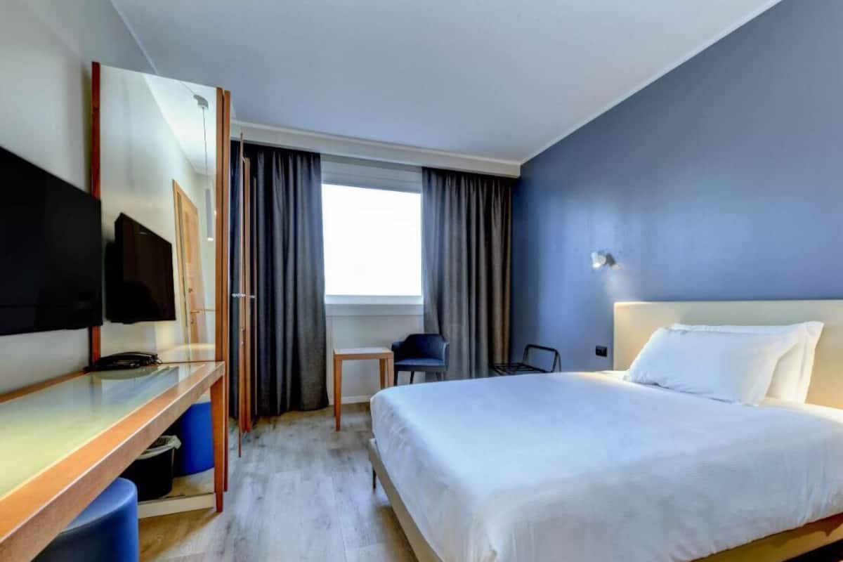 Een hotelkamer met een bed, een bureau, een stoel, een televisie en een raam met gordijnen.