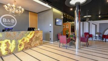 A hotel reception area with a modern design, including a marble reception desk, a chandelier, and a seating area with red chairs.