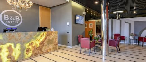A hotel reception area with a modern design, including a marble reception desk, a chandelier, and a seating area with red chairs.