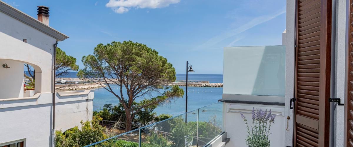 A balcony with a view of the sea, a tree, and a building with a white facade.
