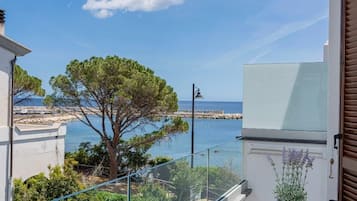 A balcony with a view of the sea, a tree, and a building with a white facade.