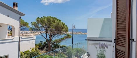 A balcony with a view of the sea, a tree, and a building with a white facade.