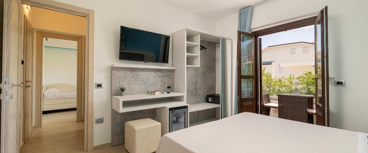 A modern hotel room with a large bed, a TV mounted on the wall, a small desk with a chair, a balcony with a wicker table, and a shower area.