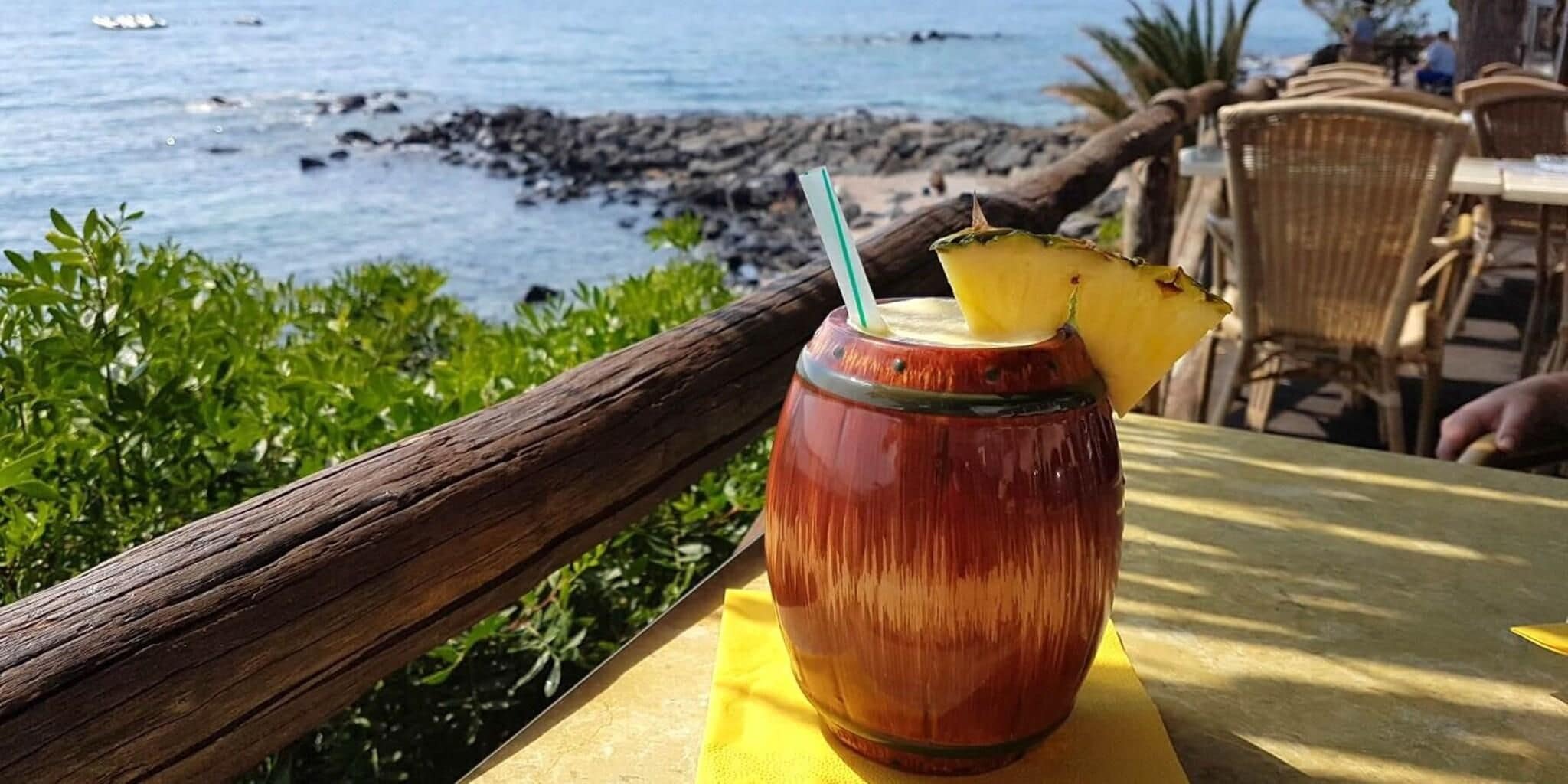 A tropical drink in a glass with a straw, placed on a yellow table with a view of the ocean and rocky shore.