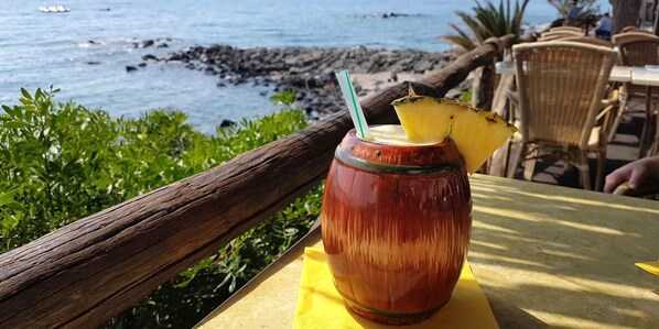 A tropical drink in a glass with a straw, placed on a yellow table with a view of the ocean and rocky shore. - Hotel La Conchiglia (Dorgali)