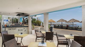 An outdoor seating area with wicker chairs and tables, a bar with a 'Cocktail' sign, and a view of the sea with thatched umbrellas.