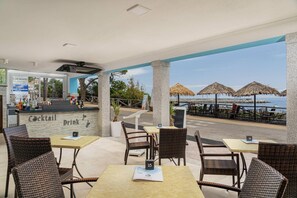 An outdoor seating area with wicker chairs and tables, a bar with a 'Cocktail' sign, and a view of the sea with thatched umbrellas. - Hotel La Conchiglia (Dorgali)