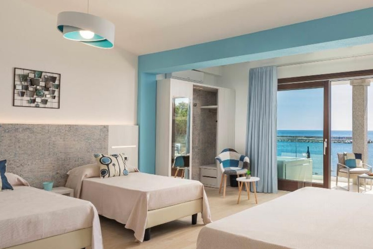 A hotel room with two beds, a balcony with a view of the sea, and a dining area with chairs and a table.