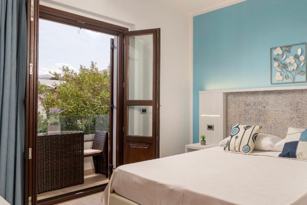 A hotel room with a bed, a nightstand, a wall-mounted lamp, a window with a view of the outdoors, and a door leading to a balcony.
