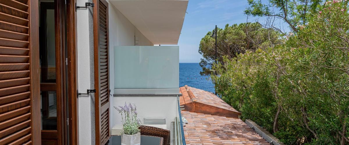 Un edificio moderno con puerta de cristal, un pequeño balcón con planta en maceta y vista al mar.