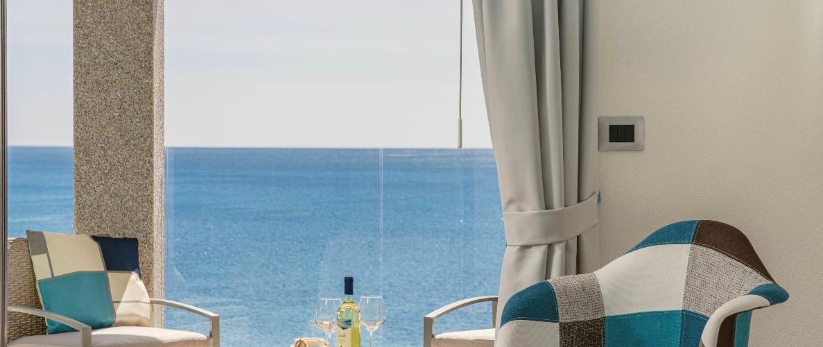 Una habitación con vista al mar, dos sillas, una mesa con una botella y una copa, y una cortina.