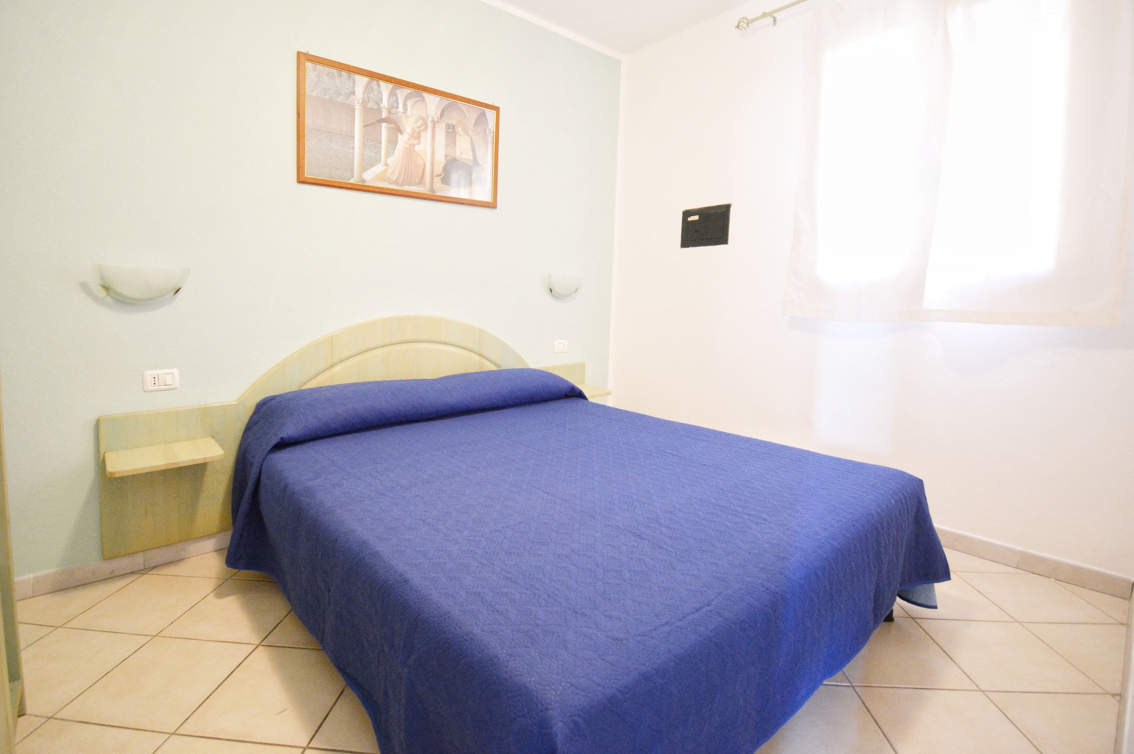 Una stanza con un letto, un comodino, una lampada a parete e un quadro incorniciato.
