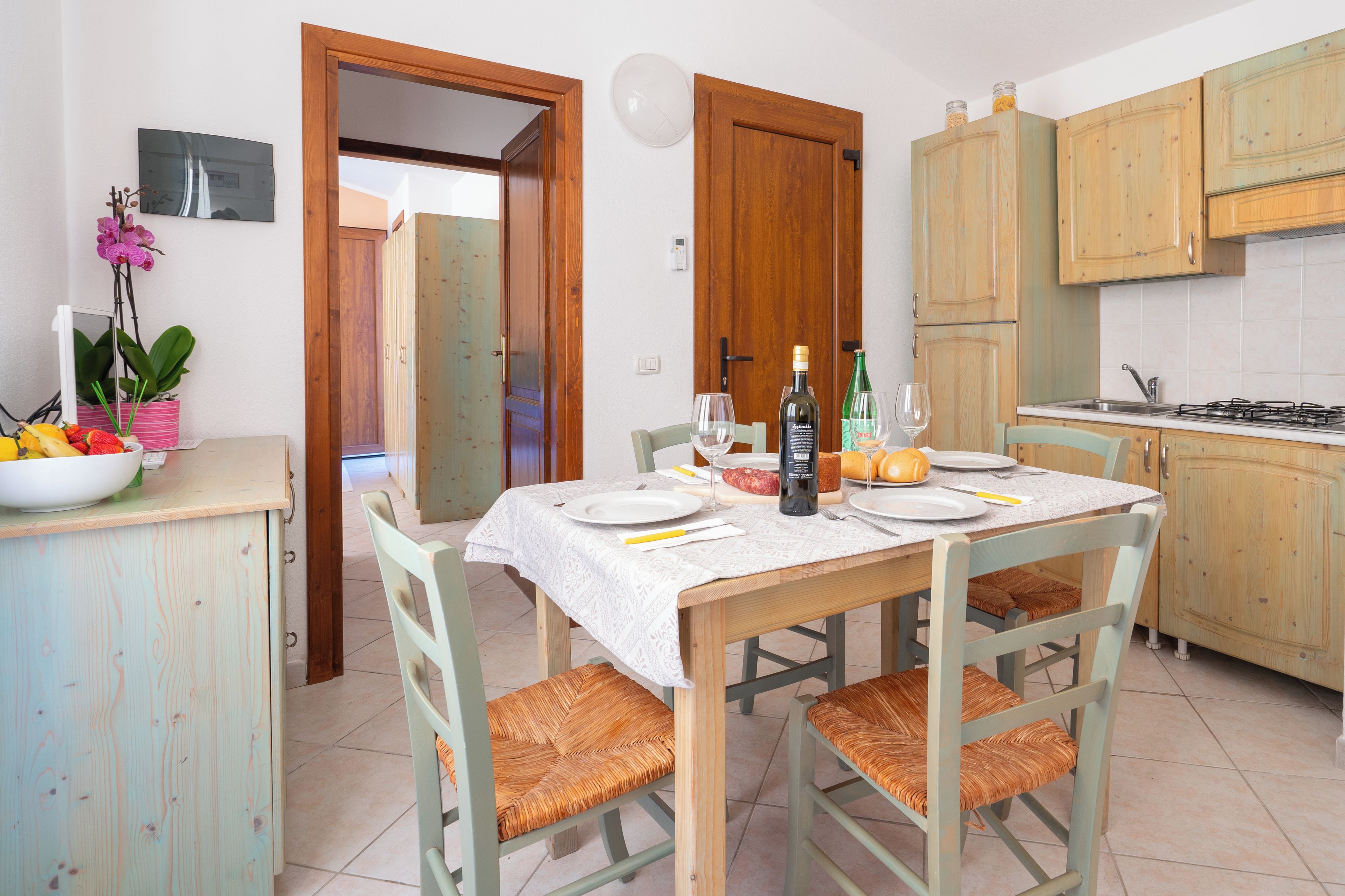A dining area with a table set for two, chairs with woven seats, a bowl of fruit, and a bottle of wine on the table.